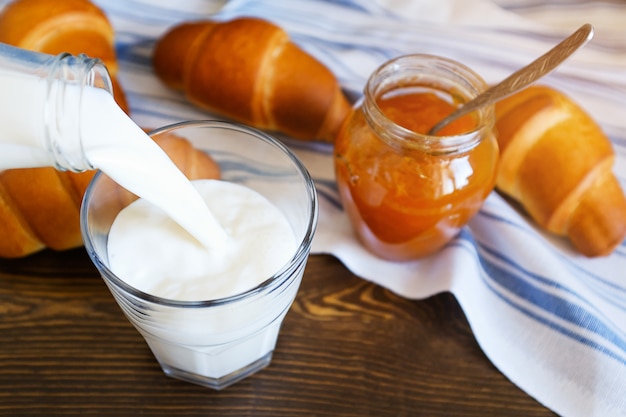 La ragazza versa il latte da una bottiglia in un bicchiere, accanto a cornetti su un piatto, marmellata. prodotti agricoli biologici Foto Premium