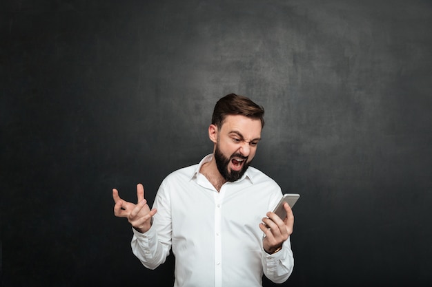 Lavoratore maschio emozionale che grida nella rabbia e nello sdegno mentre osservando sullo schermo dello smartphone d'argento sopra grigio scuro Foto Gratuite