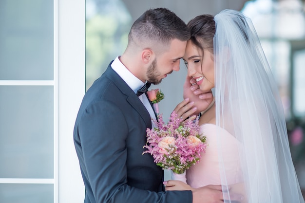 Lo sposo e la sposa in un abito rosa si tengono insieme con amore in piedi in una stanza Foto Gratuite