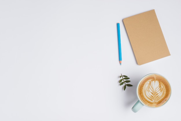 Notebook chiuso; matita colorata; foglie e tazza di caffè con latte art su sfondo bianco Foto Gratuite