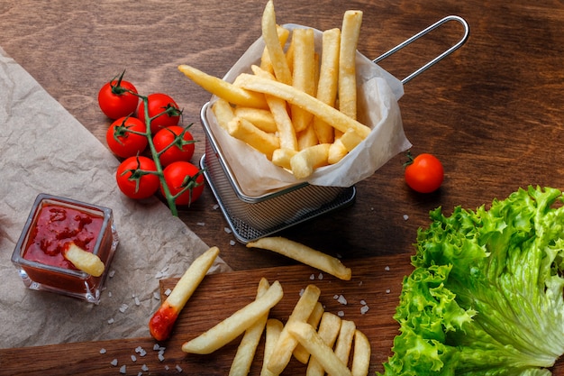 Patate fritte in una griglia con ketchup, insalata e pomodorini sul tavolo marrone in legno Foto Premium