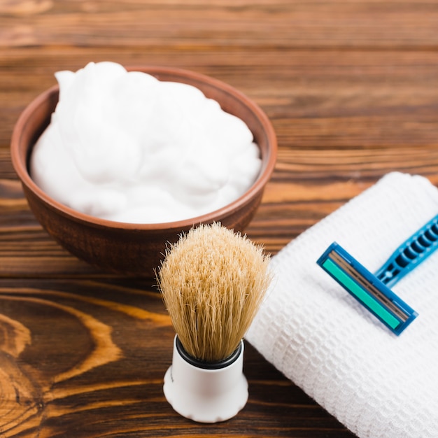 Pennello da barba sintetico; schiuma e rasoio sul tovagliolo piegato bianco sopra la scrivania in legno Foto Gratuite