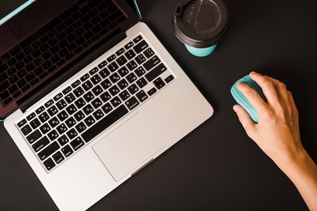 Primo piano della mano di una persona che utilizza il mouse con il computer portatile e tazza di caffè a gettare su priorità bassa nera Foto Gratuite