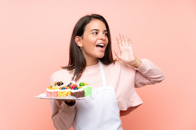 Ragazza che tiene i lotti di mini torte differenti sopra la parete isolata che grida con la bocca spalancata Foto Premium