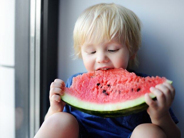 Ragazzino sveglio felice che mangia anguria che si siede sul davanzale. cibo oragnico fresco per i più piccoli Foto Premium