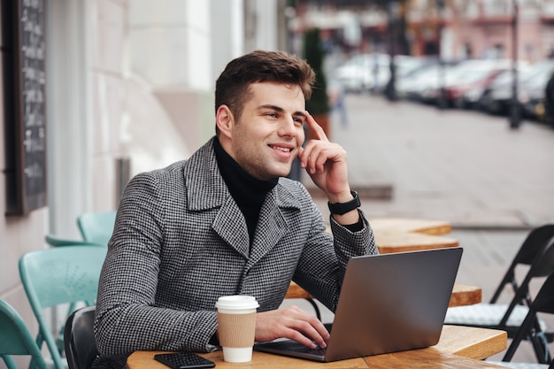 Ritratto di un ragazzo di successo che lavora con un laptop argentato in un caffè di strada, pensando al business o chiacchierando con un amico Foto Gratuite