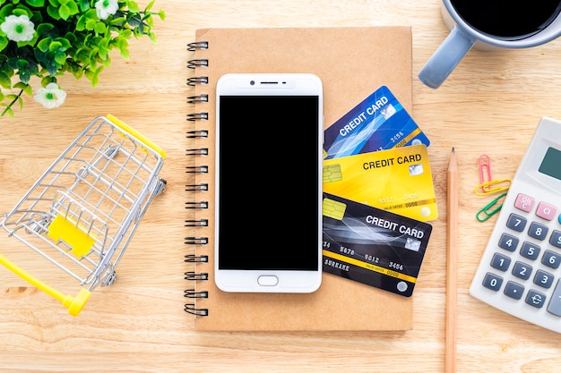Smartphone su carte di credito, taccuino, albero del vaso di fiori, carrello della spesa, calcolatrice e tazza di caffè su fondo di legno, tavolo dell'ufficio vista dall'alto bancario online Foto Premium