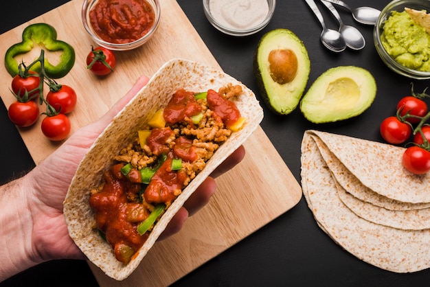 Taco della tenuta della mano vicino al tagliere fra le verdure e le salse Foto Gratuite