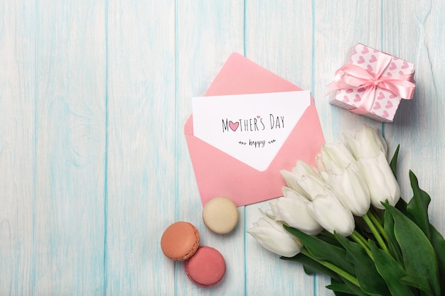 Un bouquet di tulipani bianchi con una confezione regalo, macarons, note d'amore e busta colorata su assi di legno blu. festa della mamma Foto Premium