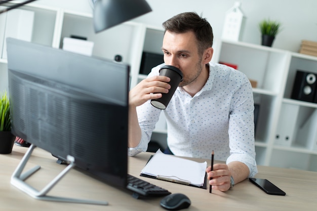 Un giovane è in piedi vicino a un tavolo in ufficio, tiene una matita in mano e beve il caffè. un giovane lavora con documenti e un computer. Foto Premium