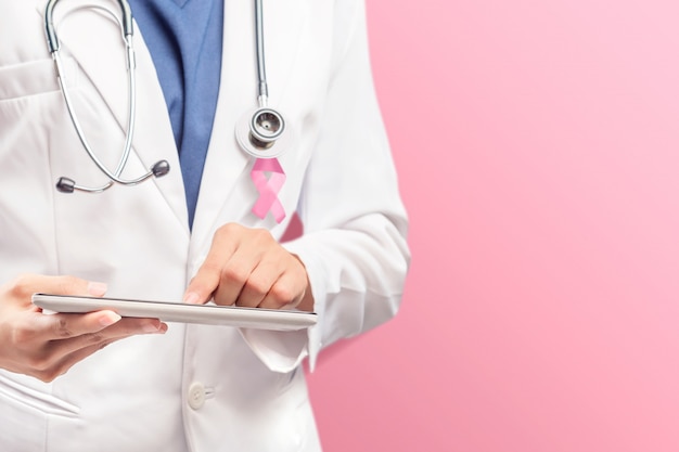 Un medico donna in un camice bianco da laboratorio con un nastro rosa e uno stetoscopio utilizzando una tavoletta con uno sfondo rosa Foto Premium