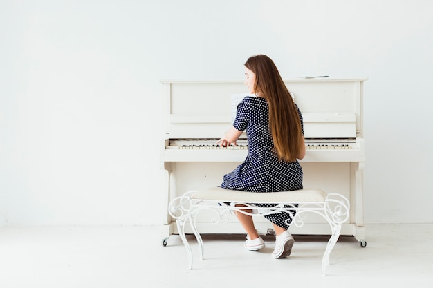 Vista posteriore di una giovane donna con i capelli lunghi, suonare il pianoforte contro il muro bianco Foto Gratuite