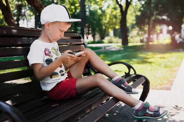 A criança senta-se no parque em um banco com um gadget. as crianças usam gadgets. retrato de um menino bonito no sol poente. um menino joga um jogo em um telefone celular. Foto Premium