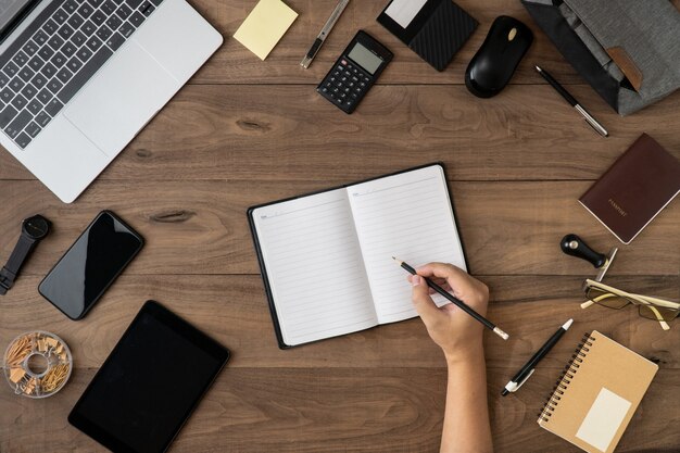 A mão direita que guarda o lápis na lista vazia do caderno com os acessórios do escritório na tabela horizontalmente leigos. Foto Premium