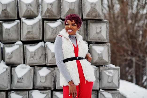 A mulher afro-americana nas calças vermelhas e no casaco branco do casaco de pele levantou no dia de inverno contra o fundo de pedra nevado. Foto Premium