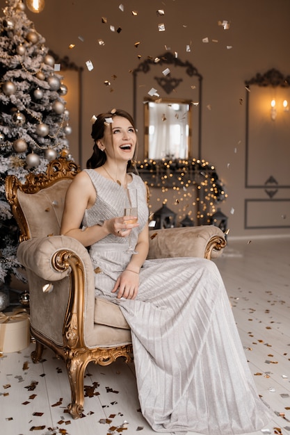 Adorável mulher de vestido prata senta-se diante de uma árvore de natal com uma taça de champanhe na mão Foto gratuita