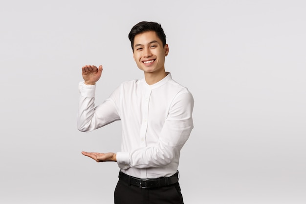 Alegre empresário asiático bonito, empregado de camisa branca, calça, mostrando uma caixa grande com expressão satisfeita, segurando o produto ou algo grande e satisfatório Foto Premium
