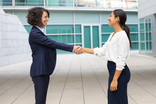Alegres colegas apertando as mãos perto do prédio de escritórios. jovens mulheres que vestem a reunião formal dos ternos ao ar livre. conceito de aperto de mão do negócio Foto gratuita