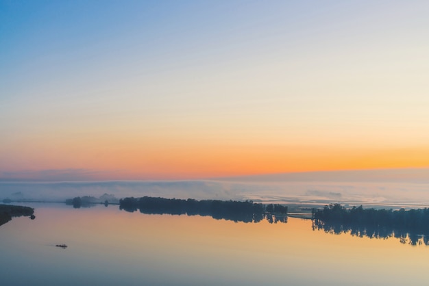 Amplo rio místico flui ao longo da costa diagonal com silhueta de árvores e névoa espessa. brilho de ouro no céu antes do amanhecer. manhã calma paisagem atmosférica de natureza majestosa em tons quentes. Foto Premium