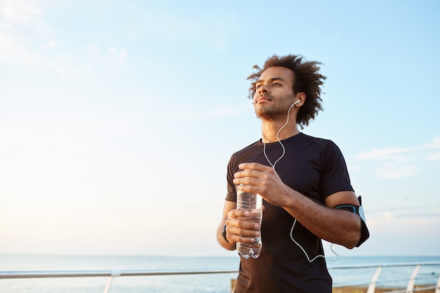 Atleta de homem bebendo água de uma garrafa de plástico após um treino de corrida difícil. esportista de pele escura olhando para o céu enquanto corre, apreciando a vista Foto gratuita