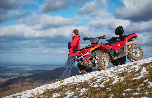 Atv moto perto do homem olha para a distância na encosta da montanha de neve na frente do céu azul nublado com espaço da cópia Foto Premium
