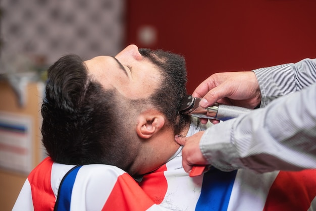 Barbeiro barbeando a barba de um homem barbudo bonito com um barbeador elétrico na barbearia