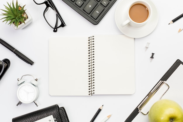 Bloco de notas em espiral rodeado com artigos de papelaria, maçã e café na mesa de escritório branco Foto gratuita