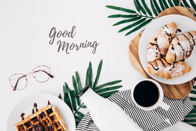 Bom dia texto com óculos; croissant cozido fresco; waffles; garrafa e xícara de café na mesa branca Foto gratuita