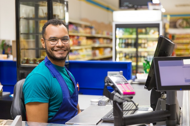 Caixa afro-americana sorridente, sentado no check-out - Grupo Muffato