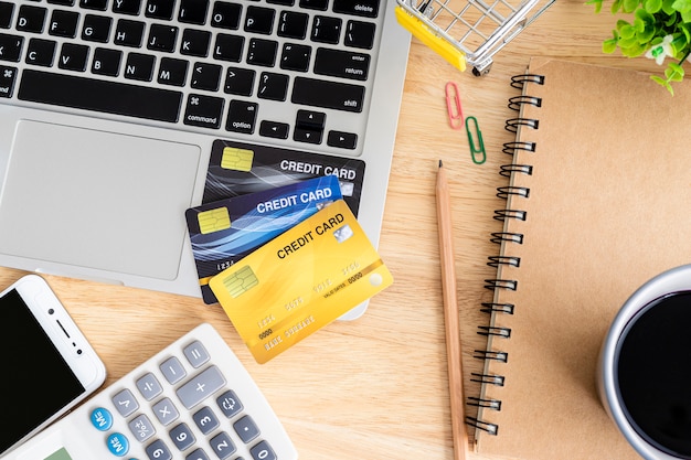 Cartão de crédito no carrinho de compras com laptop, notebook, árvore de vaso de flores, smartphone e calculadora em fundo de madeira, mesa de escritório online vista superior do banco. Foto Premium