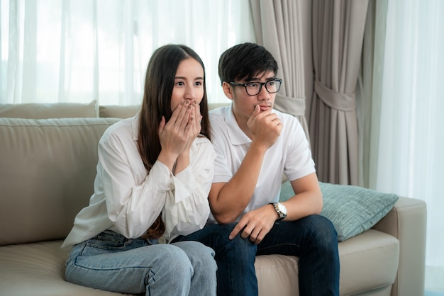 Casal asiático homem e mulher assistindo e curtindo o filme de terror tv sentado num sofá juntos na sala de estar em casa. estilo de vida familiar relaxar e conceito de recreação. Foto Premium
