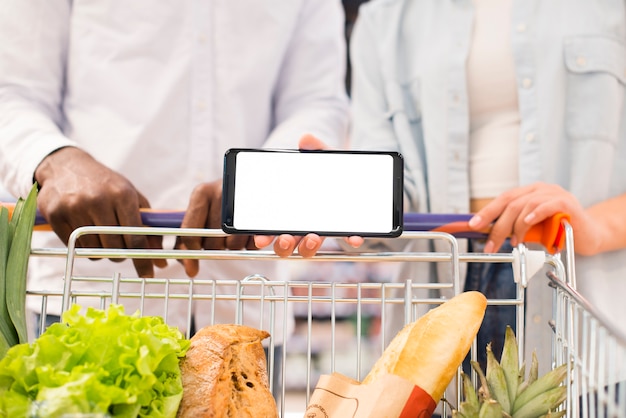 Casal sem rosto com carrinho de compras, segurando o smartphone no supermercado Foto gratuita