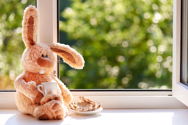 Coelho com uma xícara de café e biscoitos de manhã perto da janela aberta. bom dia e dia feliz. copie o espaço. Foto Premium