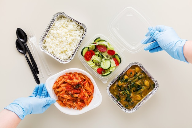 Comida para levar em descartável contaitakeaway comida em vasilhas descartáveis arroz, curry de vegetais, macarrão italiano com molho de tomate e salada fresca saudável. e mãos em luvas cirúrgicas médicas. Foto Premium