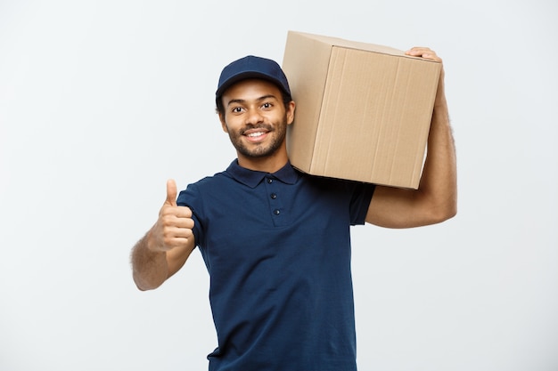 Conceito de entrega - retrato do homem de entrega feliz africano-americano em pano vermelho, segurando um pacote de caixa. isolado no fundo do estúdio cinzento. espaço de cópia. Foto gratuita