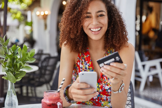 na imagem uma mulher jovem feliz e sorridente com penteado afro, usa um celular moderno e um cartão de crédito para fazer compras online | O Guia do Cartão de Crédito