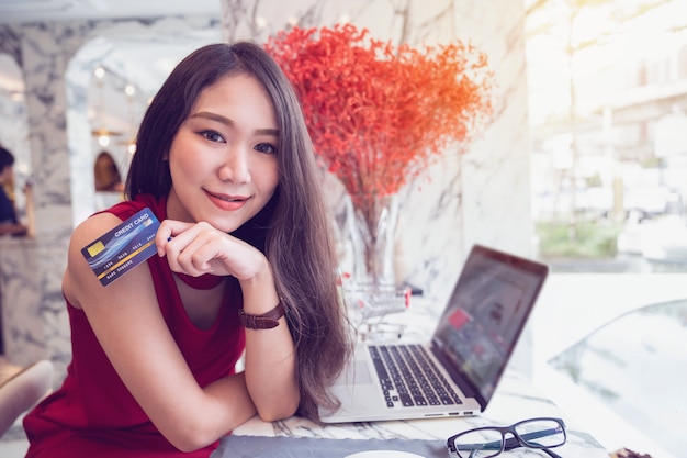 Conceitos de pagamento on-line, jovem mulher asiática sorrindo segurando o cartão de crédito nas mãos enquanto fazia compras on-line no laptop na cafeteria Foto Premium