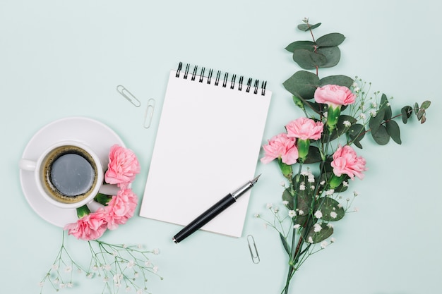 Cravos cor-de-rosa e flores de gypsophila com copo de café; bloco de notas em espiral e caneta-tinteiro em pano de fundo azul Foto gratuita