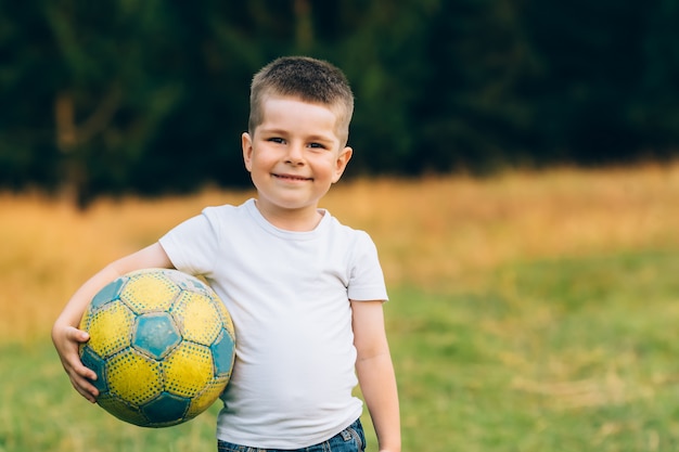 Criança com uma bola de futebol debaixo do braço no jardim de casa com fundo de grama, sorrindo Foto Premium