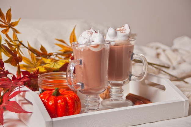 Dois copos de cacau cremoso quente com espuma na bandeja branca com folhas de outono e abóboras no fundo Foto Premium