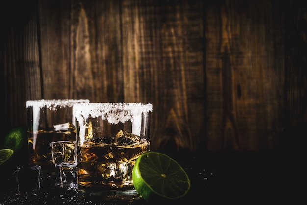 Dois copos de tequila em fundo escuro, com cubos de gelo, sal e limão Foto Premium