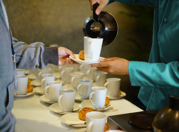 Empregado de mesa derramando quente café ou chá em copo branco e servir prato de padaria para o café tempo de pausa na festa Foto Premium