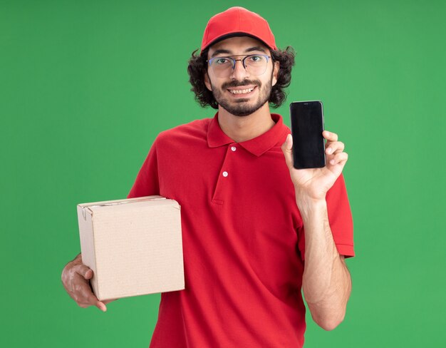 Entregador jovem sorridente com uniforme vermelho e boné de óculos segurando uma caixa de