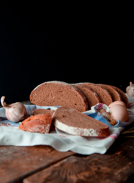 Fatias finas da opinião lateral tradicional do pão preto com farinha branca nela, em uma toalha rústica com alho e ovos, em uma mesa de cozinha de madeira. Foto gratuita