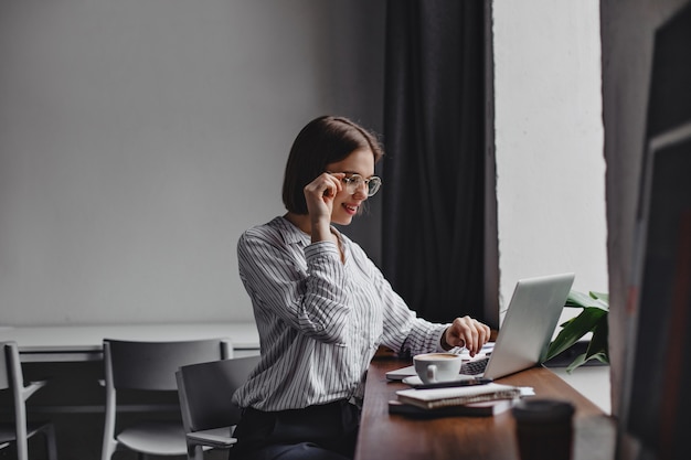 mulher de negócios de cabelos curtos de óculos e blusa branca, sentado no local de trabalho e trabalhando no laptop | Entenda o Projeto de Lei  da Reforma Tributária