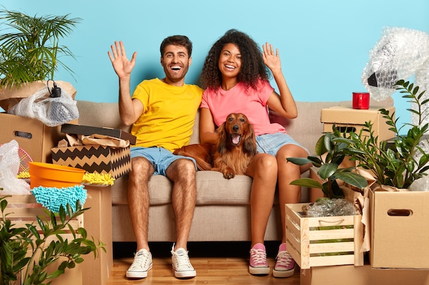 Foto interna de um casal de família feliz acenando, sente-se em um sofá confortável, o cachorro com raça está perto, comemore o dia da mudança, tenha muitas caixas com pertences para desempacotar, esteja de bom humor Foto gratuita