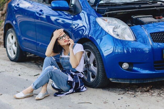 Garota está sentada ao lado de carro quebrado com um capô aberto e fala por telefone Foto Premium