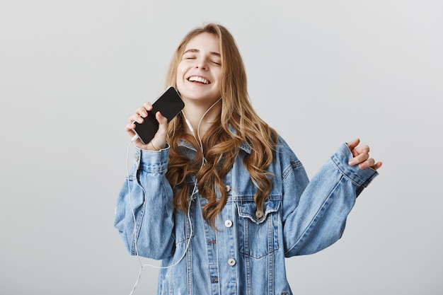 Garota loira feliz e despreocupada tocando aplicativo de karaokê no celular e cantando no smartphone com fones de ouvido | Foto Grátis