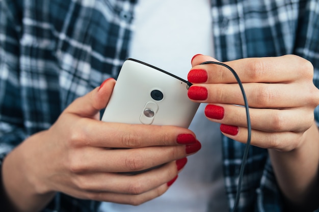 Garota na camisa xadrez, segurando um telefone inteligente nas mãos com manicure vermelho e conecta fones de ouvido Foto Premium