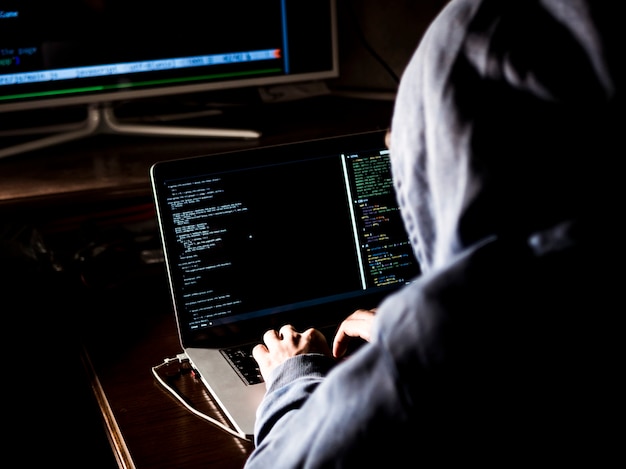 Hacker com capuz digitando código no programa usando o laptop no escuro studio Foto Premium